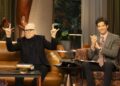 A man in black appears as a guest on a streaming series with a smiling host.