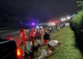 Essex Police People are sat next to the dual carriageway on the grass verge and are also walking on the A131.  It is night time with lights in the background.