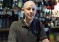 BBC Adam McClurg in a black t-shirt standing behind the bar in a pub. He is bald. There are spirits and drinks behind him.