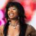 A person with long dark hair, wearing silver eye makeup and a necklace with charms, smiles against a colorful background.