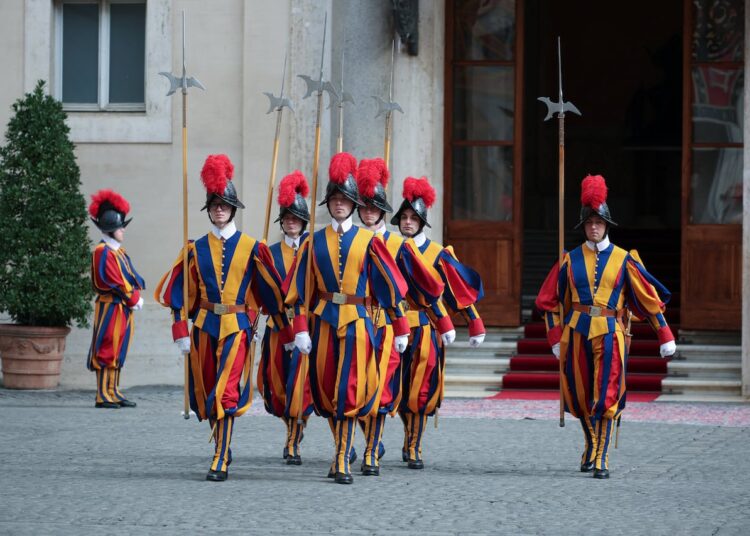 A half-dozen people in multicolored uniforms, hats and carrying ceremonial sticks walks outside.
