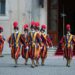 A half-dozen people in multicolored uniforms, hats and carrying ceremonial sticks walks outside.