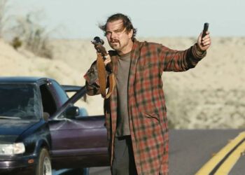 Leonardo DiCaprio as Bob holding a gun and a device in One Battle After Another.