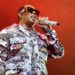 A performer in a camo jacket with "No Limit" performs on stage, holding a sparkly microphone, against a red background.