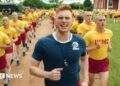 Max Parker as Sgt Sullivan, who has red hair and wears a blue sports top, shouts as he leads a training run of two rows of recruits behind him. The recruits are shaven-headed and wear yellow tops and red shorts. They are running through a field outside their training building.