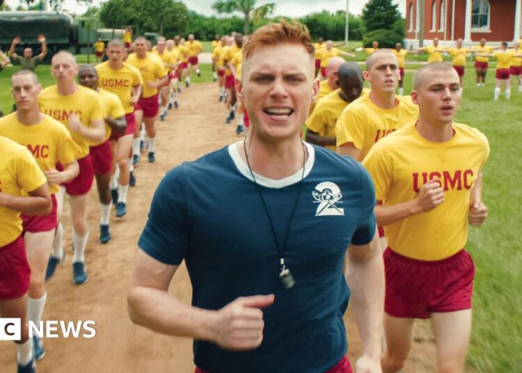 Max Parker as Sgt Sullivan, who has red hair and wears a blue sports top, shouts as he leads a training run of two rows of recruits behind him. The recruits are shaven-headed and wear yellow tops and red shorts. They are running through a field outside their training building.