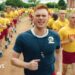 Max Parker as Sgt Sullivan, who has red hair and wears a blue sports top, shouts as he leads a training run of two rows of recruits behind him. The recruits are shaven-headed and wear yellow tops and red shorts. They are running through a field outside their training building.