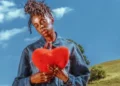 Sony Music Cat Burns holds up a cushion in the shape of a heart, while standing against a countryside landscape, in a promotional photo for her new album How To Be Human