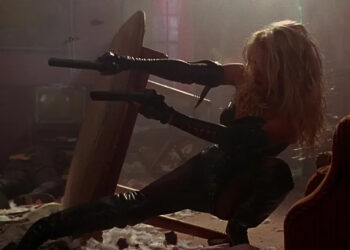 Barb firing two guns from behind a table in Barb Wire
