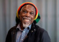 A man (Billy Ocean) is wearing a red, yellow, green and black striped hat and smiling into the camera. He has a short white beard and moustache. He is wearing a blue and white striped shirt and a black jacket. A blurred grey background is seen behind him.