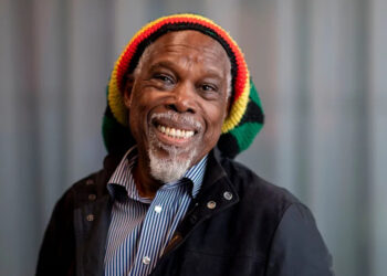 A man (Billy Ocean) is wearing a red, yellow, green and black striped hat and smiling into the camera. He has a short white beard and moustache. He is wearing a blue and white striped shirt and a black jacket. A blurred grey background is seen behind him.