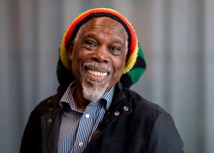 A man (Billy Ocean) is wearing a red, yellow, green and black striped hat and smiling into the camera. He has a short white beard and moustache. He is wearing a blue and white striped shirt and a black jacket. A blurred grey background is seen behind him.