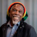 A man (Billy Ocean) is wearing a red, yellow, green and black striped hat and smiling into the camera. He has a short white beard and moustache. He is wearing a blue and white striped shirt and a black jacket. A blurred grey background is seen behind him.