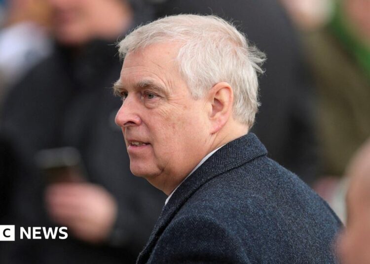 Prince Andrew wearing a dark overcoat over a white shirt, standing outdoors among a group of people. The background shows blurred figures