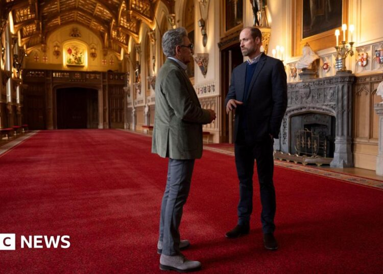 Prince William showing Eugene Levy inside Windsor Castle, they are in a large room or corridor with red carpet and ornate furnishings