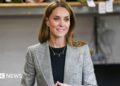 Catherine, Princess of Wales with long brown hair is wearing a grey and black blazer and black top and a gold necklace with the letters 'G, C, L' on it with small gold hoop earrings. She is smiling to the side with her hands clasped together. The background is blurred.