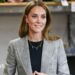 Catherine, Princess of Wales with long brown hair is wearing a grey and black blazer and black top and a gold necklace with the letters 'G, C, L' on it with small gold hoop earrings. She is smiling to the side with her hands clasped together. The background is blurred.