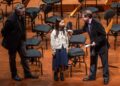 A girl speaks into a microphone held by a man in a suit, while another man stands nearby on a stage with empty chairs and music stands in the background.