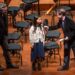 A girl speaks into a microphone held by a man in a suit, while another man stands nearby on a stage with empty chairs and music stands in the background.