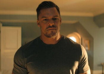 Alan Ritchson as Reacher standing in a bedroom looking down seriously.