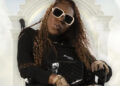 A person with long curly hair, wearing large sunglasses and patterned gloves, sits in a wheelchair against a cloudy, ornate backdrop.