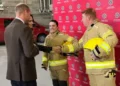 BBC A male firefighter with short ginger hair shakes hands with Catherine, Princess of Wales while a young female firefighter with dark hair stands beside them, smiling.  Prince William is standing beside his wife, talking to the recruits.  The firefighers are wearing beige safety suits and carrying bright yellow safety helmets. There is a red fire safety vehicle parked behind them.