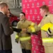 BBC A male firefighter with short ginger hair shakes hands with Catherine, Princess of Wales while a young female firefighter with dark hair stands beside them, smiling.  Prince William is standing beside his wife, talking to the recruits.  The firefighers are wearing beige safety suits and carrying bright yellow safety helmets. There is a red fire safety vehicle parked behind them.