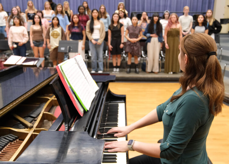 A woman dressed in green with her head turned away sits at a piano, both hands on the keyboard, and looks across the room to a group of 30 or more female students on risers, in three rows.