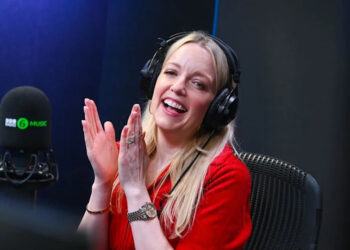 Lauren Laverne, with long blonde hair, wearing a red top and black headphones, claps her hands together, smiling, as she sits in a black chair with a microphone in front of her.