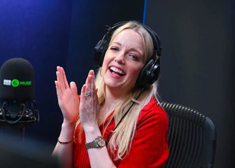 Lauren Laverne, with long blonde hair, wearing a red top and black headphones, claps her hands together, smiling, as she sits in a black chair with a microphone in front of her.