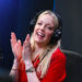 Lauren Laverne, with long blonde hair, wearing a red top and black headphones, claps her hands together, smiling, as she sits in a black chair with a microphone in front of her.