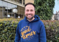 James Murden, co-founder of Good Times Live wearing a blue hoodie with the Good Times logo on it in mustard. He has dark brown hair and a short beard. Behind him is a hedge, with buildings behind that.