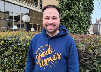 James Murden, co-founder of Good Times Live wearing a blue hoodie with the Good Times logo on it in mustard. He has dark brown hair and a short beard. Behind him is a hedge, with buildings behind that.