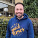 James Murden, co-founder of Good Times Live wearing a blue hoodie with the Good Times logo on it in mustard. He has dark brown hair and a short beard. Behind him is a hedge, with buildings behind that.