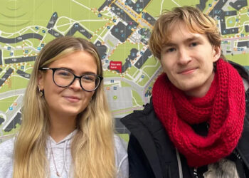 Grace Mahon and Charles Murphy, are standing side by side in front of a large, detailed campus map. Grace is wearing glasses and a light-coloured hoodie, while Charles is dressed in a black jacket with a bright red scarf. The map behind them shows buildings and pathways, indicating their location within the university grounds. Grace has long blonde hair and Charles has shorter blonde hair.