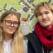 Grace Mahon and Charles Murphy, are standing side by side in front of a large, detailed campus map. Grace is wearing glasses and a light-coloured hoodie, while Charles is dressed in a black jacket with a bright red scarf. The map behind them shows buildings and pathways, indicating their location within the university grounds. Grace has long blonde hair and Charles has shorter blonde hair.