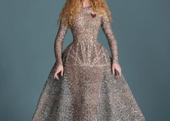 A woman with orange hair wears a structured silver dress which is tight at the top and flares at her waist. It has a red heart.