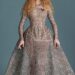 A woman with orange hair wears a structured silver dress which is tight at the top and flares at her waist. It has a red heart.