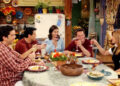 Five people sitting around a table, each holding a glass and smiling, as if toasting. Plates of food and autumn-themed decorations are on the table