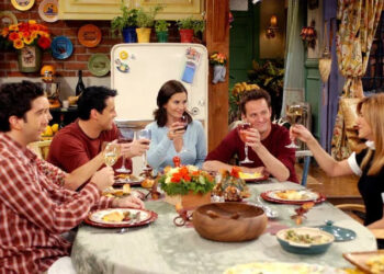 Five people sitting around a table, each holding a glass and smiling, as if toasting. Plates of food and autumn-themed decorations are on the table