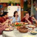 Five people sitting around a table, each holding a glass and smiling, as if toasting. Plates of food and autumn-themed decorations are on the table