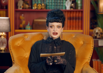 A woman in a black dress and lace gloves sits in a yellow armchair, holding an envelope.