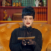 A woman in a black dress and lace gloves sits in a yellow armchair, holding an envelope.