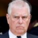 Prince Andrew, who has short white hair, wears a formal black suit, white shirt, and black tie. He stands against a dark background with another person partially visible behind him. His expression is serious and composed.