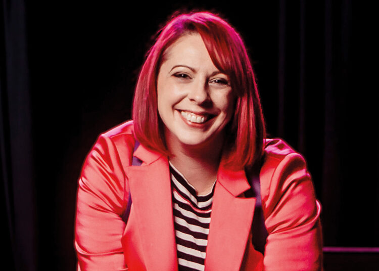 A woman (Carrie Davenport) in a bright pink blazer sits confidently with a camera, ready to capture moments in a dimly lit venue