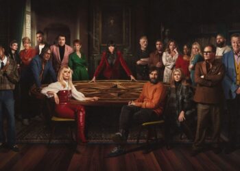The cast of The Celebrity Traitors stand around a circular table wearing poker faces whilst the show's host, Claudia Winkleman, stands centre.