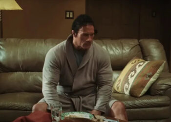 Dwayne Johnson as Mark Kerr sitting in a robe on the couch in The Smashing Machine