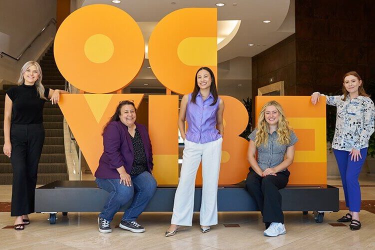 Five Titan alumni who work for the OCVIBE development pose in front of a display.