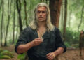 Henry Cavill as Geralt with long grey hair and cuts all over his face. He holds a silver pendant. He's wearing an open-buttoned green shirt with sleeves rolled up. He's in a forest.