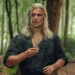 Henry Cavill as Geralt with long grey hair and cuts all over his face. He holds a silver pendant. He's wearing an open-buttoned green shirt with sleeves rolled up. He's in a forest.
